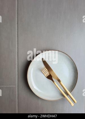 Assiette en céramique de couleur naturelle avec rebord marron et couteau de couleur or avec fourchette sur fond gris, assiette avec couverts Banque D'Images