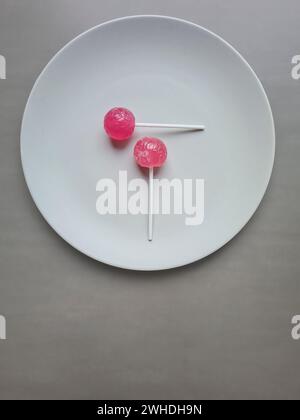 Deux sucettes rouge clair aromatisées à la fraise, sucettes avec une tige blanche reposent sur une assiette Banque D'Images