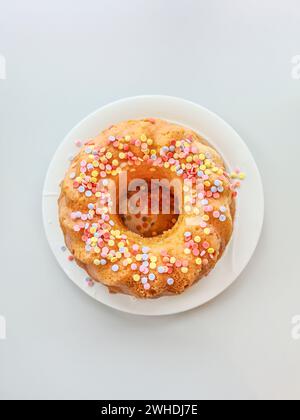 Gâteau éponge rond avec des saupoudrages colorés et la garniture se dresse sur une assiette blanche devant un fond blanc Banque D'Images