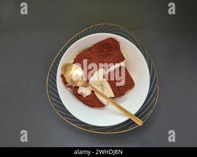 Une portion fraîche de tiramisu dans un bol blanc avec une cuillère à dessert sur un fond sombre Banque D'Images