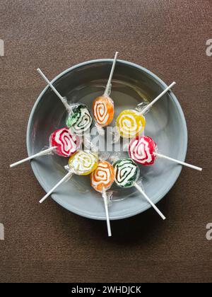 Huit sucettes colorées sont encore enveloppées dans une feuille de plastique en cercle dans un bol gris Banque D'Images