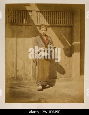 La photographie montre une jeune femme, pleine longueur, debout, face à l'avant, portant une théière et un petit plateau avec des tasses à thé ; elle peut porter un tablier autour de sa taille et est peut-être serveuse dans un salon de thé. Vue extérieure du bâtiment en arrière-plan..... Japon colorié à la main avec aquarelle tirage photographique vers les années 1870 Banque D'Images