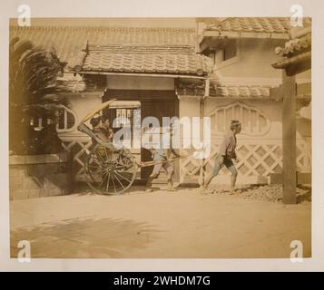 La photographie montre deux femmes avec un grand parasol chevauchant dans un pousse-pousse tiré par un homme avec un deuxième homme marchant à proximité. Vue extérieure du bâtiment avec toit en tuiles à l'arrière-plan....Japon coloré à la main avec aquarelle Photographic Print vers les années 1870 Banque D'Images