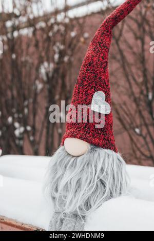 Nain avec symbole de coeur sur le banc dans la neige. Cadeau romantique. Concept de la Saint-Valentin. GNOME avec coeur et chapeau rouge dans la neige. Fond d'amour. Banque D'Images