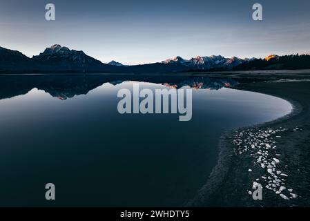 Panorama alpin avec reflet dans le Forggensee partiellement drainé et hivernal près de Füssen dans le Allgäu. Alpenglow dans la première lumière du soleil. Au premier plan, des pierres sur le lit gelé du lac. Banque D'Images