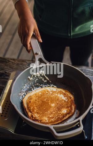 femme faisant des crêpes dans la poêle à frire, tenant la poêle avec la main droite sur le poêle de camp extérieur Banque D'Images