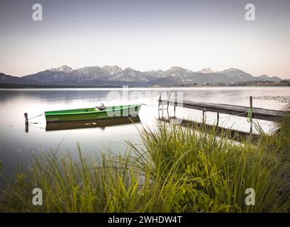 Bateau à rames vert amarré à une jetée en bois sur le lac Hopfensee avec vue sur les Alpes de Allgäu. Roseaux et herbes au premier plan. Le bateau et la jetée se reflètent dans l'eau calme. Prise le matin. Hopfen am See, Hopfensee, East Allgäu, Allgäu, Bavière Banque D'Images