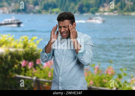 Jeune homme stressé souffrant de terribles maux de tête debout à l'extérieur. Homme avec mal de tête sur fond de mer. Banque D'Images