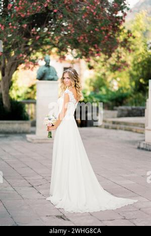 Mariée dans une robe blanche avec un bouquet se tient dans le jardin contre la toile de fond d'un arbre à fleurs Banque D'Images