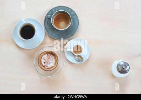 Diverses boissons au café comme l'espresso, le cappuccino, le lungo noir et avec de la crème et un distributeur de sucre sur une table en bois clair, panor Banque D'Images