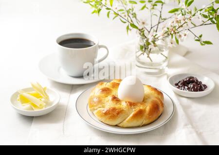 Pain de levure de Pâques tressé en forme de nid avec un œuf, du café, du beurre et de la confiture comme petit déjeuner festif du dimanche sur une table blanche, copie Banque D'Images