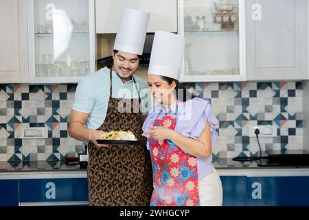 Heureux couple indien habillé en uniforme de cuisinier portant un tablier préparant des aliments savoureux dans la cuisine. Montrer les compétences de cuisine Banque D'Images