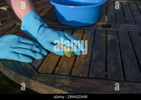 Mains dans des gants en caoutchouc bleu nettoyant une table de jardin en bois altérée au début du printemps ou la saison extérieure, espace de copie Banque D'Images
