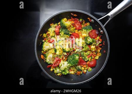 Repas de légumes sautés à base de brocoli, poivron, oignon et tomate dans une poêle sur la cuisinière noire, cuisine végétarienne Banque D'Images