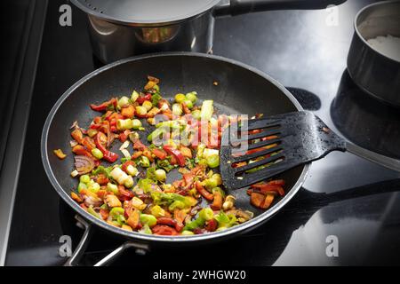 Cuire les légumes sautés comme le poivron, l'oignon et le poireau avec des épices dans une poêle noire sur la cuisinière avec d'autres casseroles, cuisiner Banque D'Images