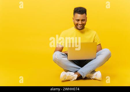 Homme indien souriant utilisant un ordinateur portable assis sur le sol dans la pose détendue, isolé sur fond jaune, étudiant masculin serein insouciant étudiant au loin, freelance travaillant à distance Banque D'Images