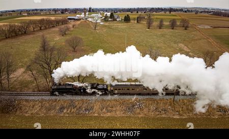 Vue aérienne latérale d'un train de passagers à vapeur approchant, voyageant à travers des terres agricoles ouvertes, soufflant beaucoup de fumée blanche Banque D'Images