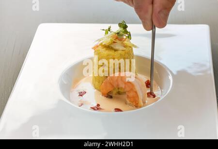 Main avec une pince à épiler garnissant un plat gastronomique de risotto au safran avec garniture aux herbes, une grosse crevette et une sauce aux crustacés dans un mod Banque D'Images