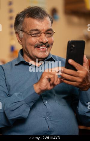 Plan vertical, homme âgé souriant heureux à l'aide d'un téléphone mobile avec des lunettes à la maison - concept de vie de retraite, contenu des médias sociaux et cyberespace Banque D'Images