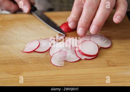 Les mains coupent les radis en fines tranches avec un couteau de cuisine sur une planche à découper en bois pour une salade fraîche, espace copie, focus sélectionné Banque D'Images