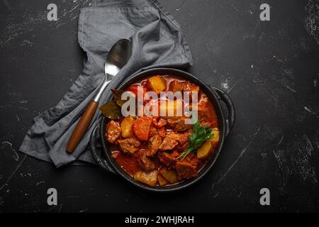 Ragoût de viande de bœuf avec pommes de terre, carottes et sauce délicieuse dans une casserole noire avec feuilles de Laurier et cuillère sur noir rustique foncé Banque D'Images