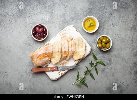 Ciabatta fraîche tranchée sur planche de cuisine à découper, olives vertes et brunes, huile d'olive au romarin, branches d'olivier sur con gris Banque D'Images