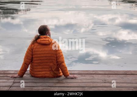 Homme solitaire en veste orange de randonnée en duvet assis sur la jetée en bois regardant le reflet du ciel dans l'eau Banque D'Images