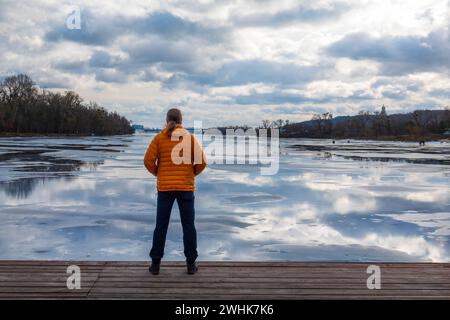 Homme solitaire en veste en duvet orange debout sur une jetée en bois sur la rive de la rivière regardant les reflets du ciel dans l'eau et la glace. Banque D'Images