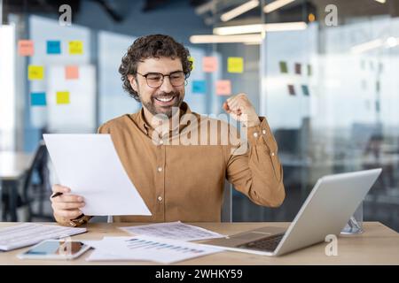 Financier comptable prospère à l'intérieur du bureau sur le lieu de travail, homme heureux célèbre la victoire, travail de papier de succès, homme d'affaires tient le rapport dans les mains, satisfait des résultats de la réalisation. Banque D'Images