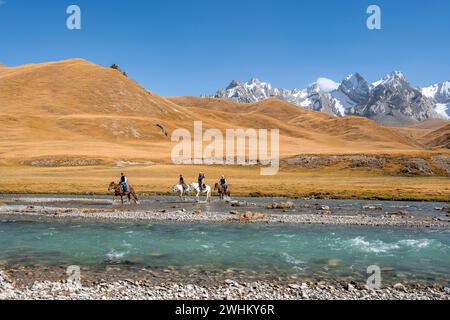 Cavaliers chevauchant la rivière en face du paysage de montagne avec des prairies jaunes, la rivière Kol Suu et les sommets de montagne, Keltan Mountains, Sary Beles Banque D'Images