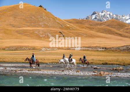 Cavaliers chevauchant la rivière en face du paysage de montagne avec des prairies jaunes, la rivière Kol Suu et les sommets de montagne, Keltan Mountains, Sary Beles Banque D'Images