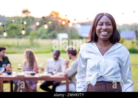 Belle jeune femme noire avec de longs cheveux droits dans une blouse blanche avec un groupe d'amis en arrière-plan ayant un outdoo Banque D'Images