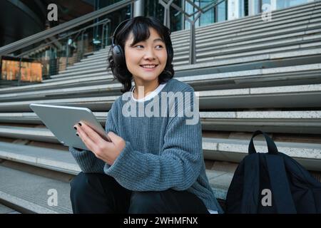 Étudiant chinois souriant assis dans les escaliers avec tablette, dessine de l'art numérique, projet de conception graphique pour le travail freelance, écoute de la musique i. Banque D'Images