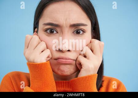 Gros plan de la femme asiatique bouleversante en colère, coulant et froncant les sourcils, tenant les mains sur les joues, se sent offensé, se tient au-dessus du fond bleu Banque D'Images