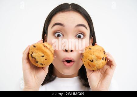 Gros plan portrait de la femme coréenne, semble surpris, montre deux cupcakes, fond blanc Banque D'Images