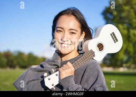 Portrait de belle fille souriante avec ukulele, femme asiatique avec instrument de musique posant à l'extérieur dans le parc vert Banque D'Images