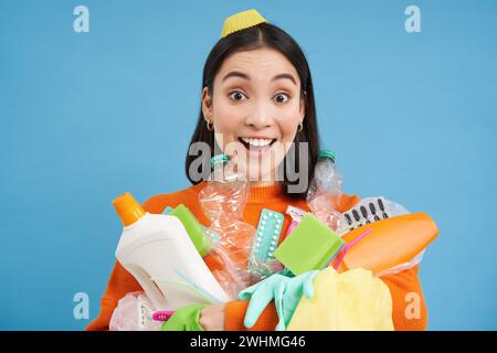 Portrait de femme coréenne enthousiaste, tenant des bouteilles en plastique vides, triant les ordures, souriant et paraissant heureuse, isolée sur bl Banque D'Images