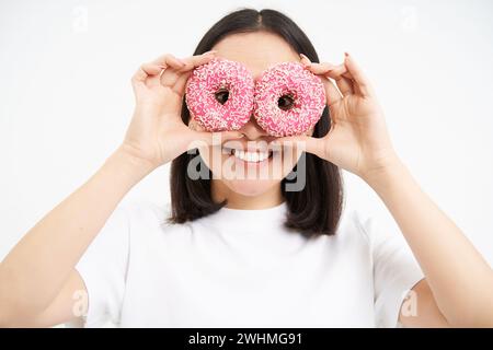 Image de jeune femme drôle, fait des verres avec deux beignets glacés roses, regardant à travers les trous de donnut et souriant, isolé sur Banque D'Images