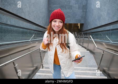 Portrait redhead fille touriste, monte les escaliers avec smartphone, suit l'itinéraire sur l'application de téléphone mobile, tient sac à dos et sourit Banque D'Images