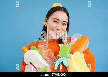 Portrait de femme coréenne enthousiaste, tenant des bouteilles en plastique vides, triant les ordures, souriant et paraissant heureuse, isolée sur bl Banque D'Images