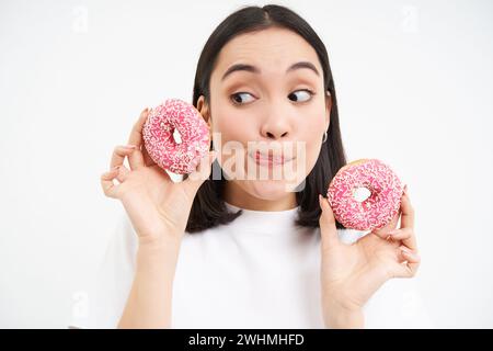 Portrait de drôle de femme asiatique, tentée de manger savoureux dougnut glacé, tenant deux donnuts et souriant excité, isolé sur b blanc Banque D'Images