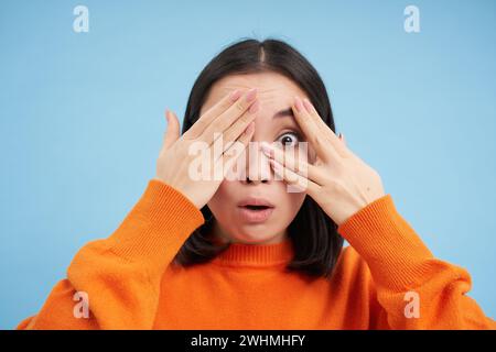 Gros plan portrait d'une jeune fille coréenne attend la surprise, ferme les yeux et regarde à travers les doigts avec le sourire excité, se tient au-dessus de bl Banque D'Images