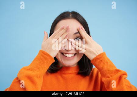 Gros plan portrait d'une jeune fille coréenne attend la surprise, ferme les yeux et regarde à travers les doigts avec le sourire excité, se tient au-dessus de bl Banque D'Images