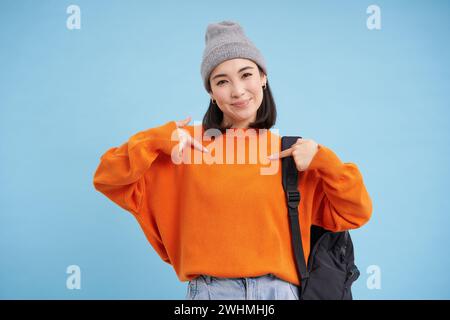 Une femme coréenne élégante se pointe sur elle-même, se tient dans un chapeau chaud et tient le sac à dos sur l'épaule, sur fond bleu Banque D'Images