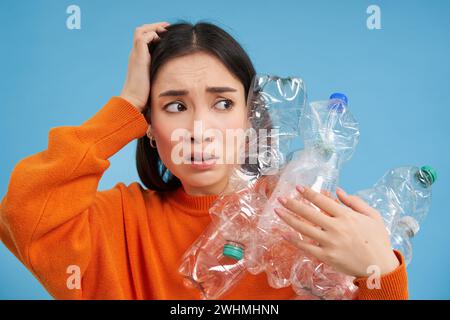 Portrait d'une fille asiatique au visage effrayé, tenant des piles de bouteilles en plastique pour le recyclage et regarde vers la droite avec emo choqué Banque D'Images