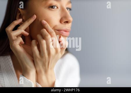 Attrayante jeune femme bouleversée en peignoir blanc examine les boutons sur son visage de près. Peau problématique sur le visage, acné. Portra Banque D'Images