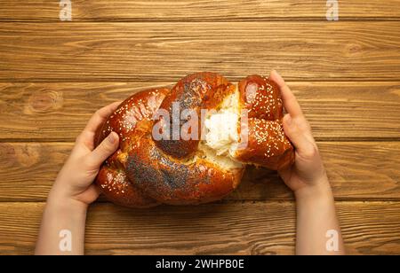 Mains féminines arrachant un morceau de pain Challah fraîchement cuit recouvert de pavot et de graines de sésame, vue de dessus sur bois rustique b Banque D'Images