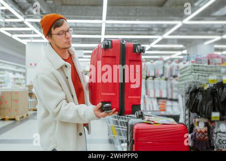 Un homme dans un supermarché examine une valise rouge pour un voyage ou un voyage d'affaires, vérifie soigneusement la qualité du produit, le choix. Banque D'Images