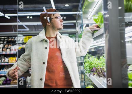 Un jeune homme attentif dans une tenue d'hiver élégante choisit la laitue fraîche sur une étagère réfrigérée dans la section des légumes d'un supermarché. Banque D'Images