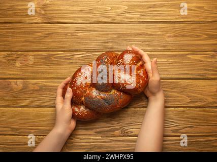 Mains féminines tenant du pain Challah fraîchement cuit recouvert de graines de pavot et de sésame, vue de dessus sur fond de bois rustique, trad Banque D'Images